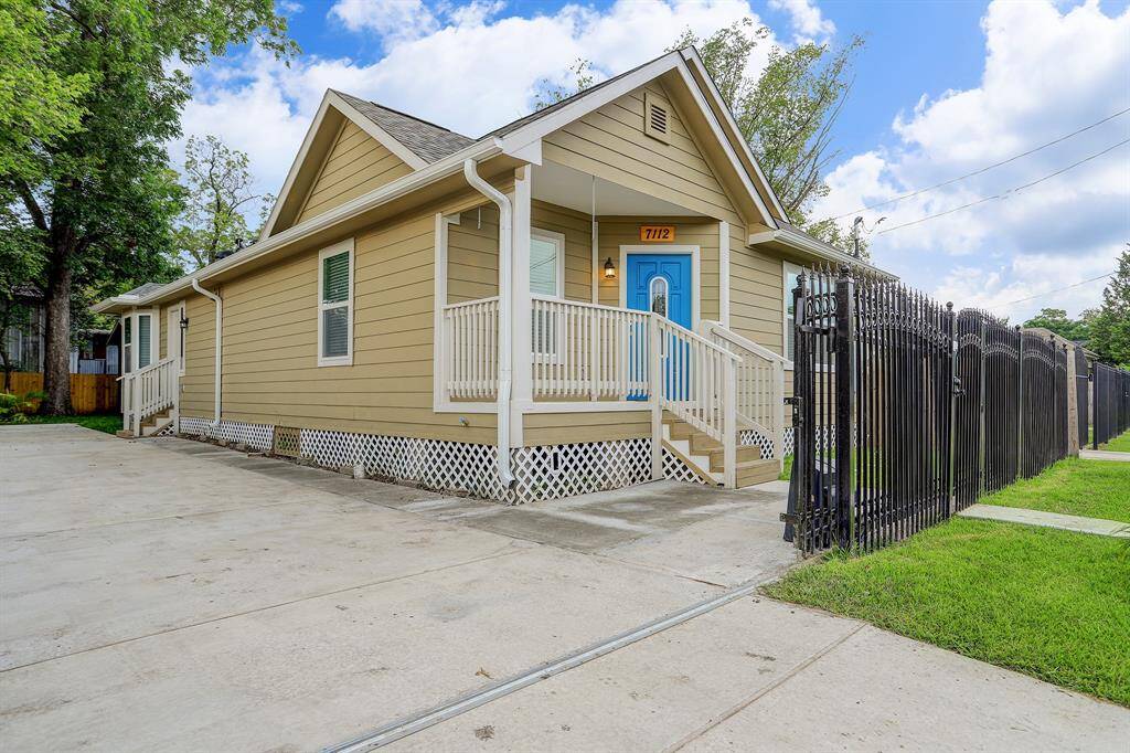 Charming single-story home featuring a welcoming front porch, beige siding, and a bright blue door, complemented by a secure black iron fence and a spacious concrete driveway.