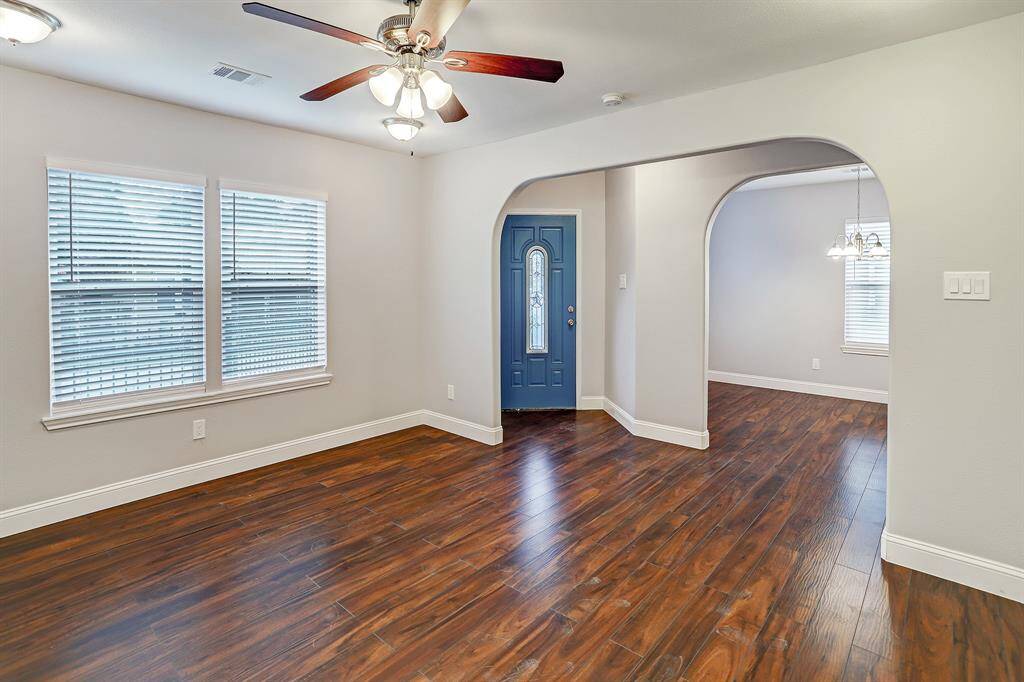 This is a spacious, well-lit room featuring polished floors, a ceiling fan with lighting, two windows with blinds, and a neutral color scheme. An arched doorway leads to the adjoining dinning room, and there's a stylish blue front door.