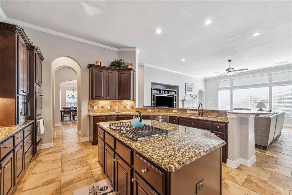 Spacious open-plan kitchen and living area with granite countertops, dark wood cabinets, and tile flooring. Features a large island with a gas cooktop, adjacent dining area, and ample natural light from large windows. Ideal for entertaining.