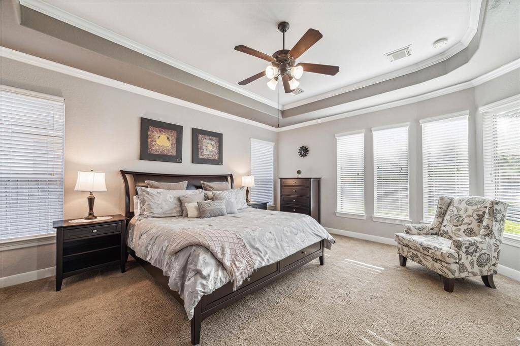 Bright and spacious bedroom with elegant tray ceiling and ceiling fan. Large windows allow natural light to flood the room, creating a warm and inviting atmosphere.