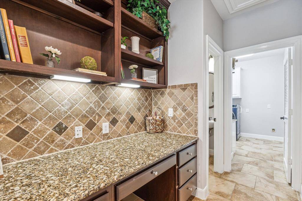 A cozy workspace nook with granite countertops and a stone tile backsplash. It features dark wood shelving for additional storage and decorative display. The area is well-lit with under-cabinet lighting.
