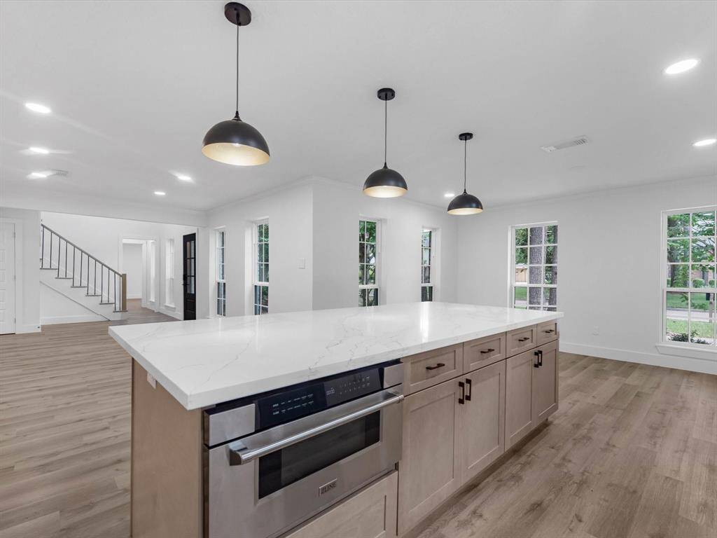 This spacious, modern kitchen features a large island with a marble countertop, pendant lighting, and ample cabinetry. The open floor plan leads to a bright living area with hardwood floors and large windows, offering a welcoming and airy atmosphere.