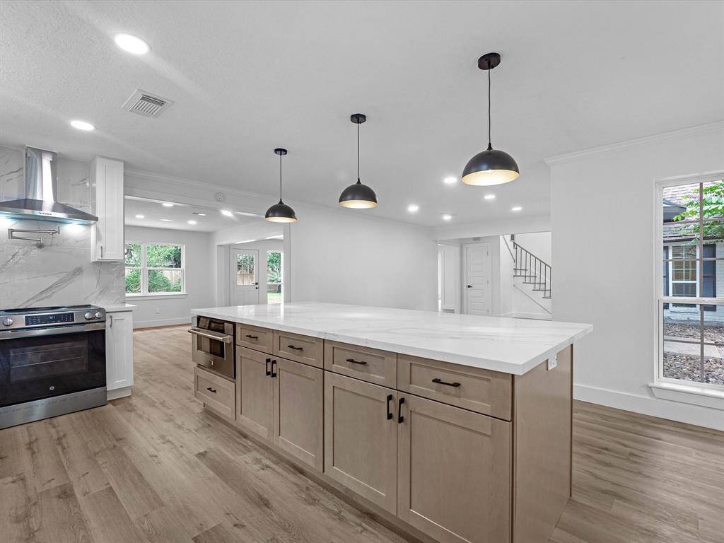 Modern kitchen with an open layout, featuring a spacious island with pendant lighting. It has wood flooring, sleek cabinetry, and stainless steel appliances. Large windows provide natural light, and there's a view of the adjacent living area.