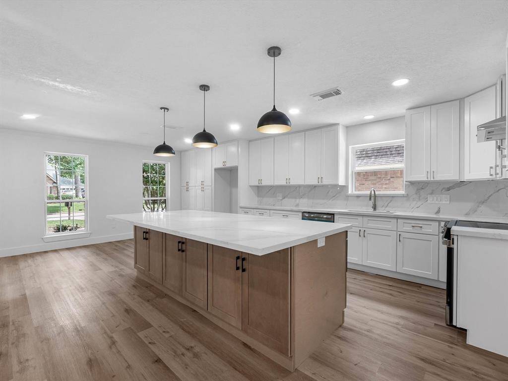 This photo showcases a spacious, modern kitchen with a large central island featuring a sleek white countertop. The kitchen is equipped with white cabinetry and stainless steel appliances, complemented by wood flooring and pendant lighting. Large windows provide ample natural light.