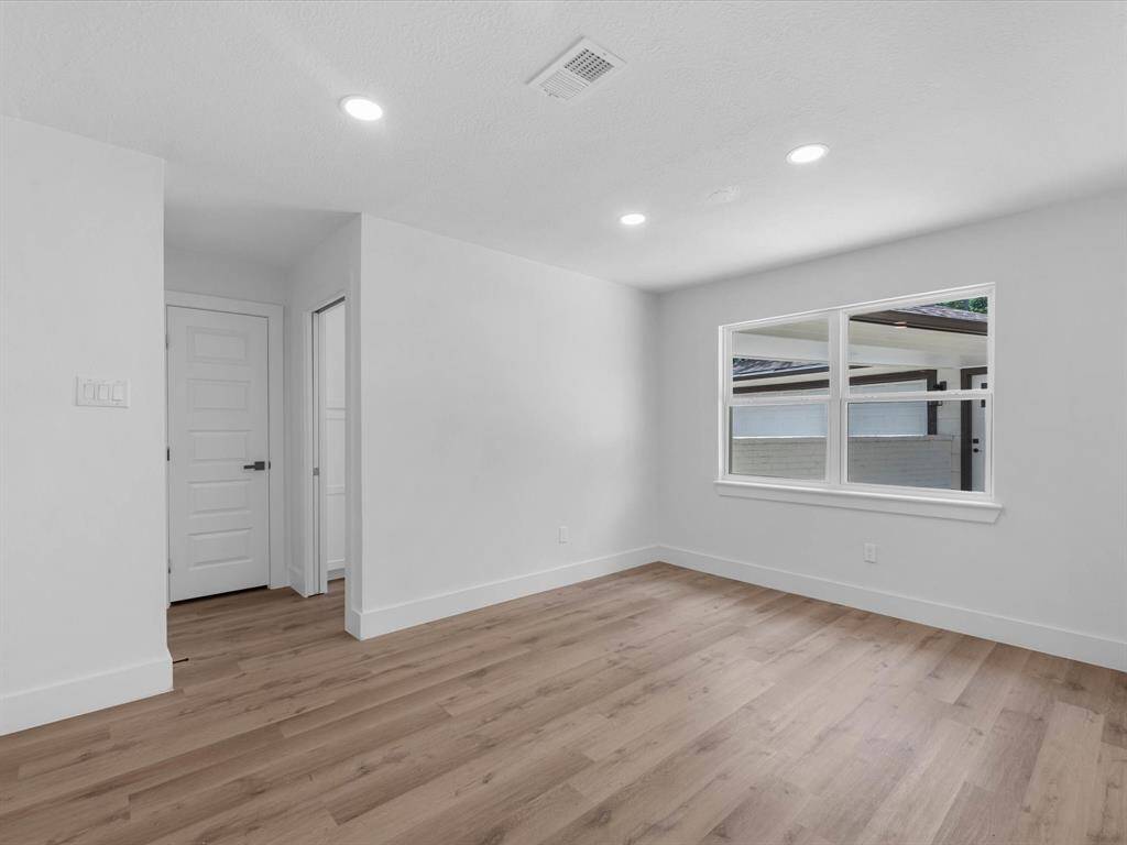 This photo showcases a bright, unfurnished room with light wood flooring, recessed lighting, and a large window offering natural light. The room features a white door and a clean, modern aesthetic.