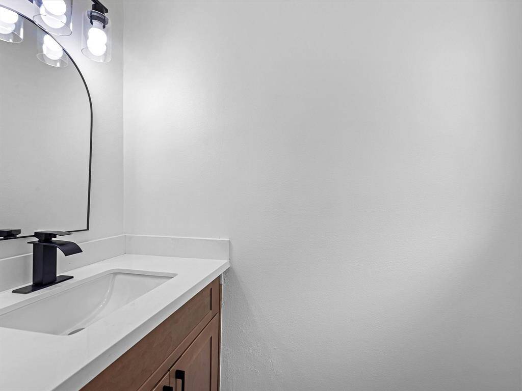 This photo shows a modern bathroom with a sleek white countertop and a rectangular sink. The vanity features a dark faucet and wood cabinetry, complemented by a large, stylish mirror with contemporary lighting above. The overall look is clean and minimalistic.