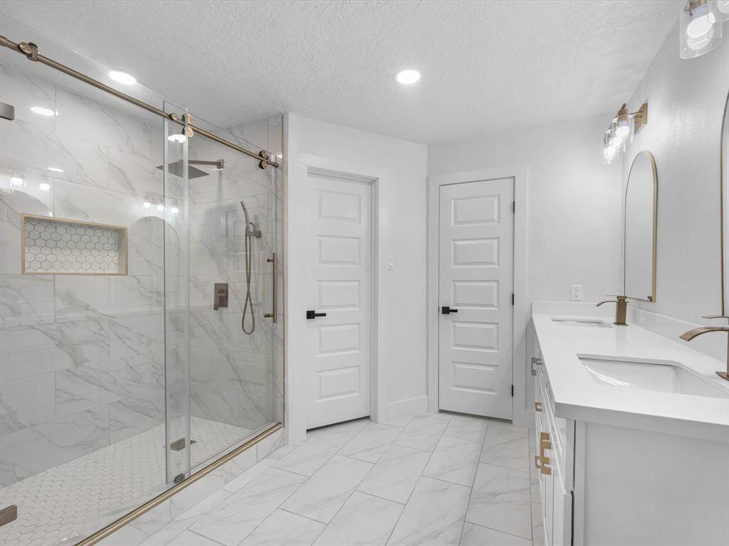 This bathroom features a spacious glass-enclosed shower with marble-style tiles and a built-in niche. It includes a double vanity with modern fixtures, sleek white cabinetry, and elegant lighting, creating a bright and contemporary space.