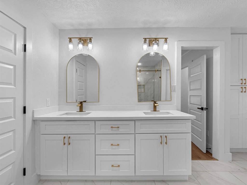This modern bathroom features a double vanity with sleek white cabinetry and gold fixtures. It has two stylish mirrors, elegant lighting, and a clean, contemporary design with a neutral color palette.
