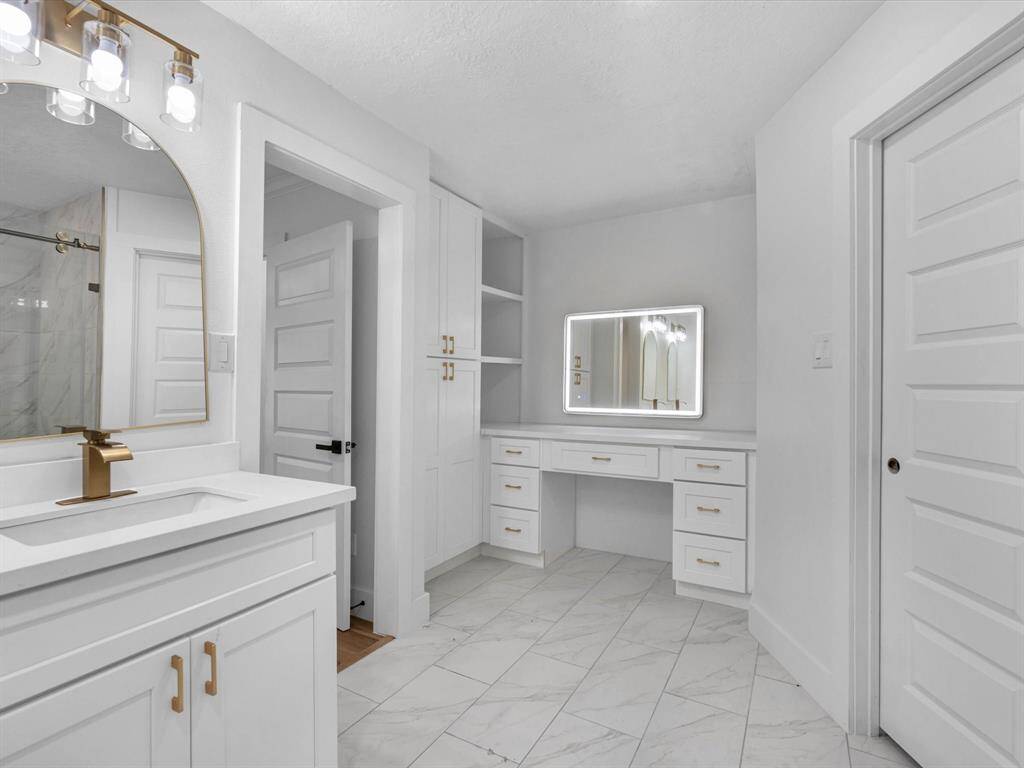 This bathroom features a modern design with white cabinetry, gold hardware, and a sleek marble-like tile floor. It includes a spacious vanity with a large illuminated mirror, ample storage, and elegant lighting fixtures for a bright and airy feel.