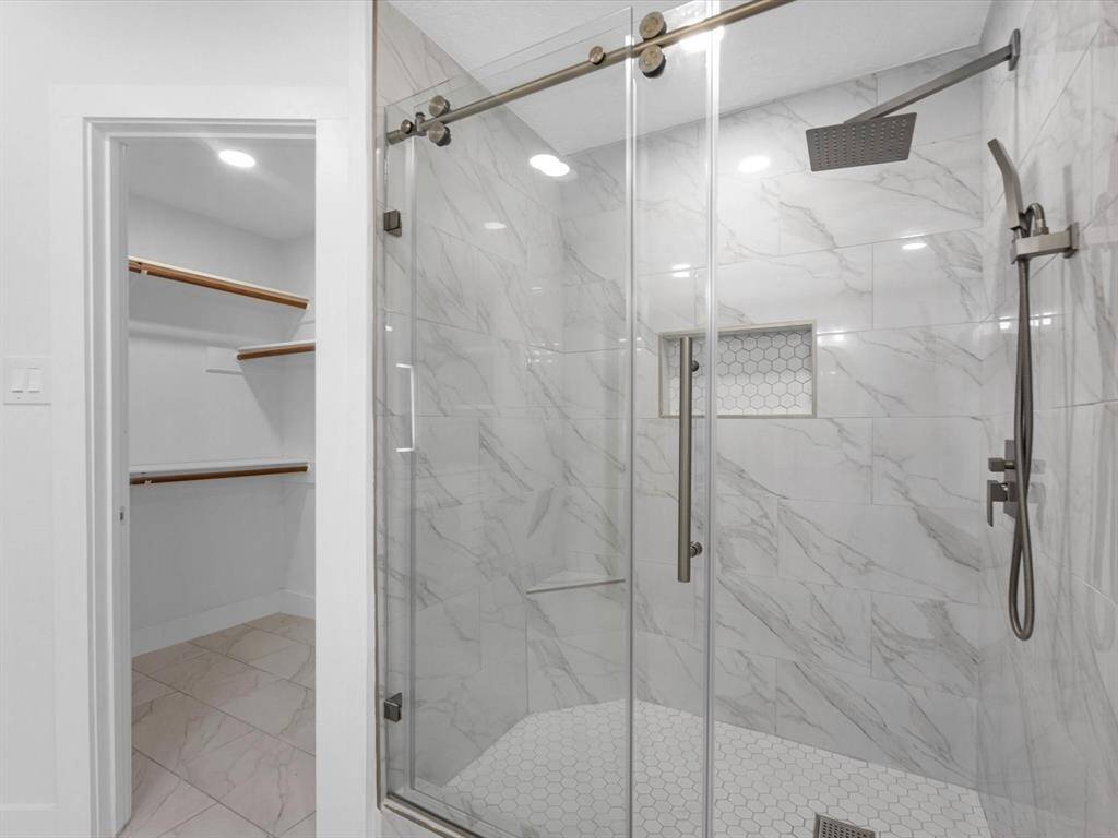 This photo showcases a modern bathroom with a spacious walk-in shower featuring elegant marble-style tiles, a rainfall showerhead, and sliding glass doors. Adjacent is a well-lit walk-in closet with ample shelving and hanging space.
