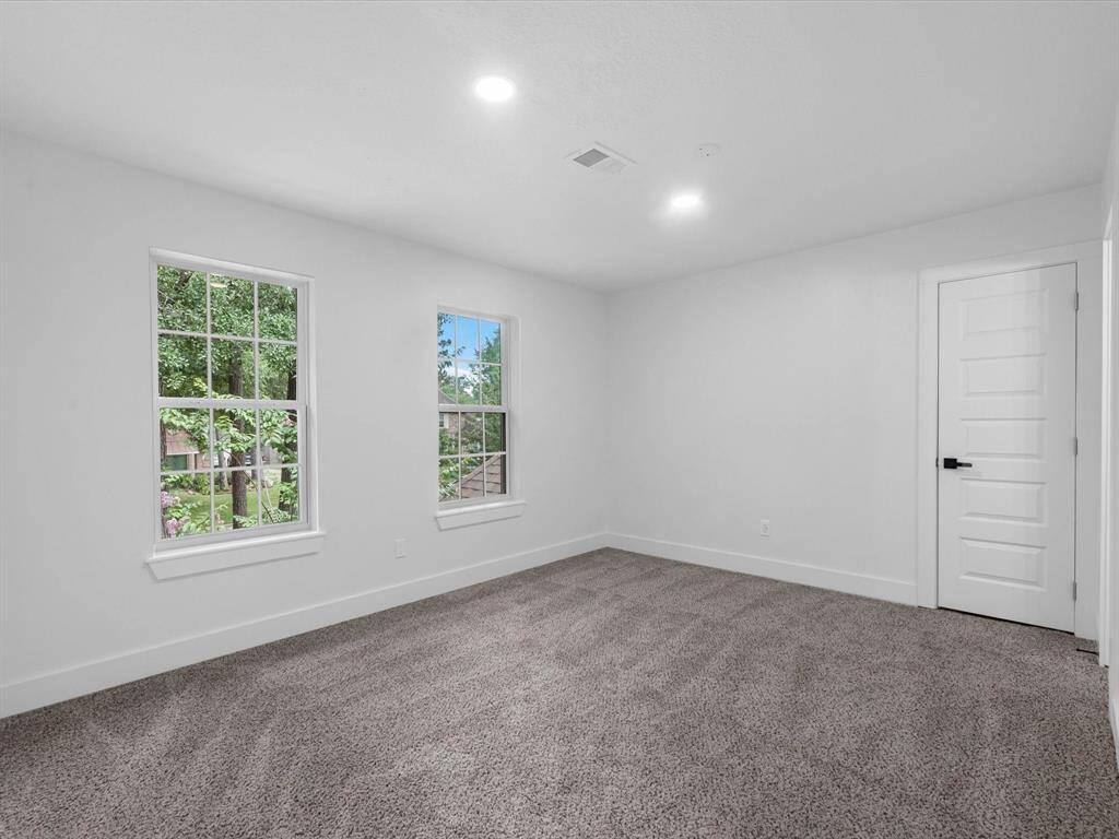 This photo shows a bright, spacious bedroom with neutral carpeting and white walls. It features two large windows offering natural light and outdoor views, along with a modern door. Ideal for creating a cozy and inviting personal space.