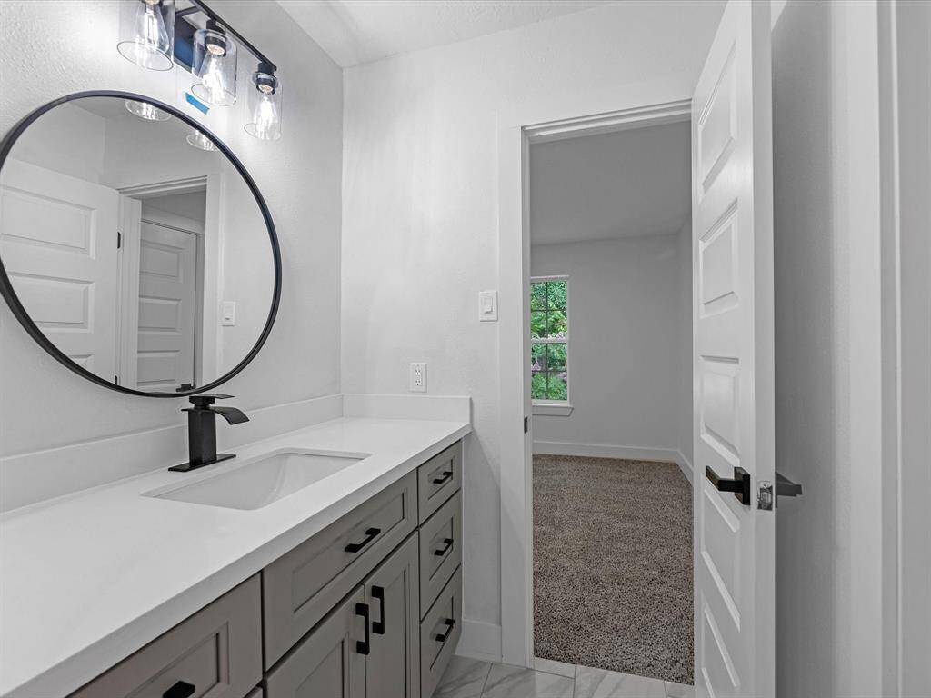 This photo shows a modern bathroom with a sleek vanity featuring a white countertop and black fixtures. A round mirror and stylish light fixtures enhance the space. A door leads to a carpeted room with a window.