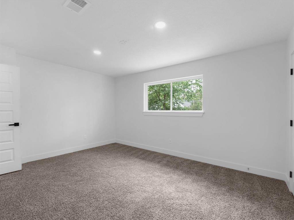 This room is spacious with neutral walls and plush carpet flooring. It features a large window allowing ample natural light and views of greenery. Recessed lighting and a modern door complete the look, offering a cozy and versatile space.