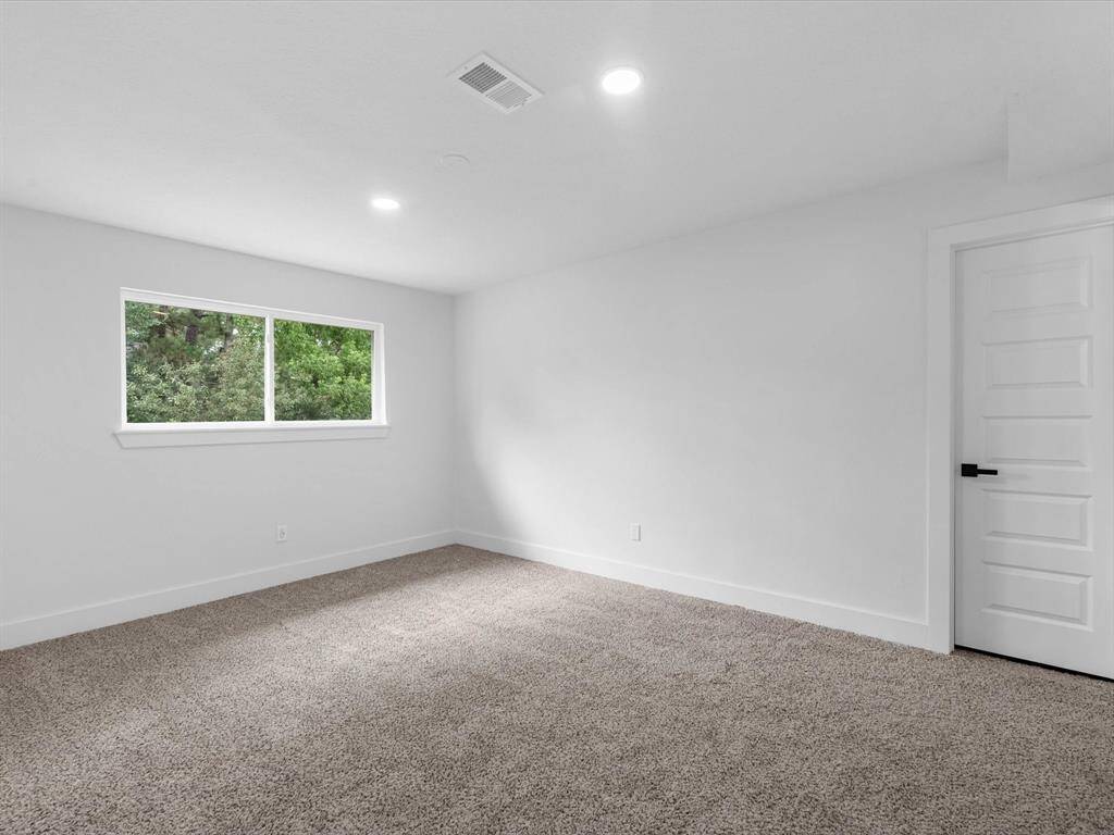 This is a bright, spacious room with neutral walls and carpeted flooring. It features a large window providing natural light and a modern white door. Ideal for a bedroom or home office.