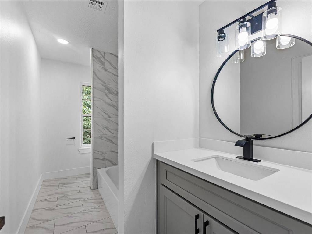 Modern bathroom with a sleek vanity featuring a round mirror and stylish lighting. The space includes a tub with elegant tilework and a window for natural light.