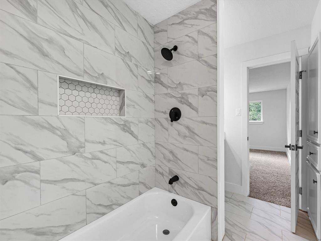 This bathroom features a modern bathtub with elegant marble-style wall tiles and matte black fixtures. It includes a hexagonal tile niche for storage and an adjoining bedroom view.