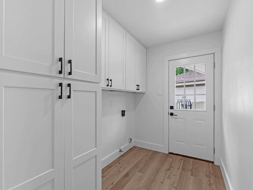 This photo shows a bright laundry room or mudroom with white cabinetry, wood flooring, and a door leading outside. The space is clean, modern, and functional with ample storage.