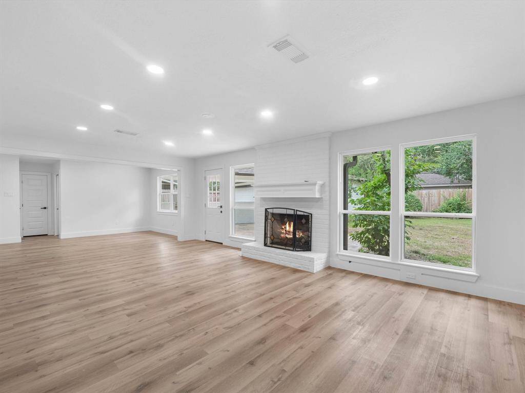 This spacious living room features light wood flooring, a cozy white brick fireplace, and large windows offering plenty of natural light and views of the backyard. The open layout provides ample space for various seating arrangements.