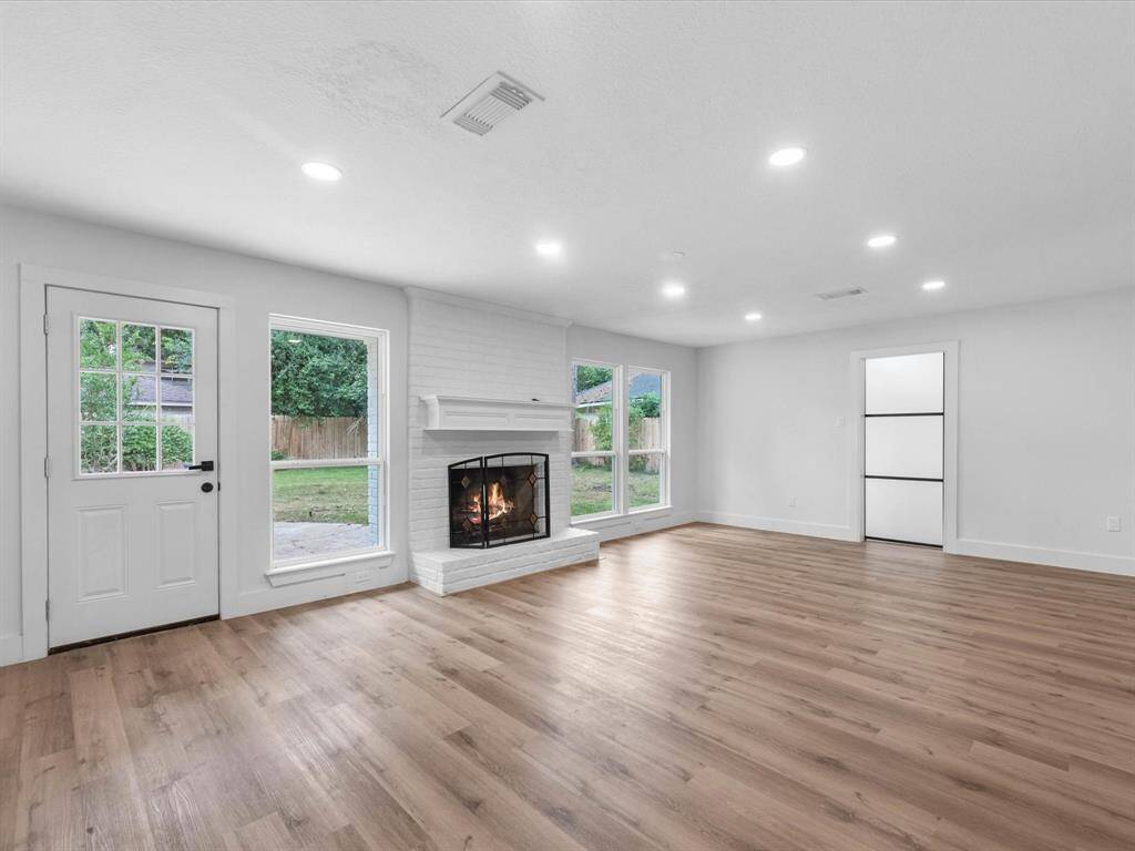 This spacious living room features a cozy fireplace, elegant wood flooring, and ample natural light from large windows. The room offers a bright, airy feel with recessed lighting and a view of the backyard through a glass door.