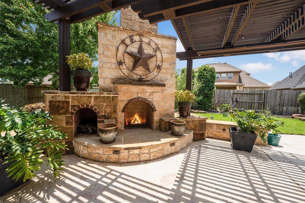 Experience the charm of a custom wood-burning fireplace with a stone surround, nestled under a lighted pergola, complemented by a soothing fountain and elegant landscaping.