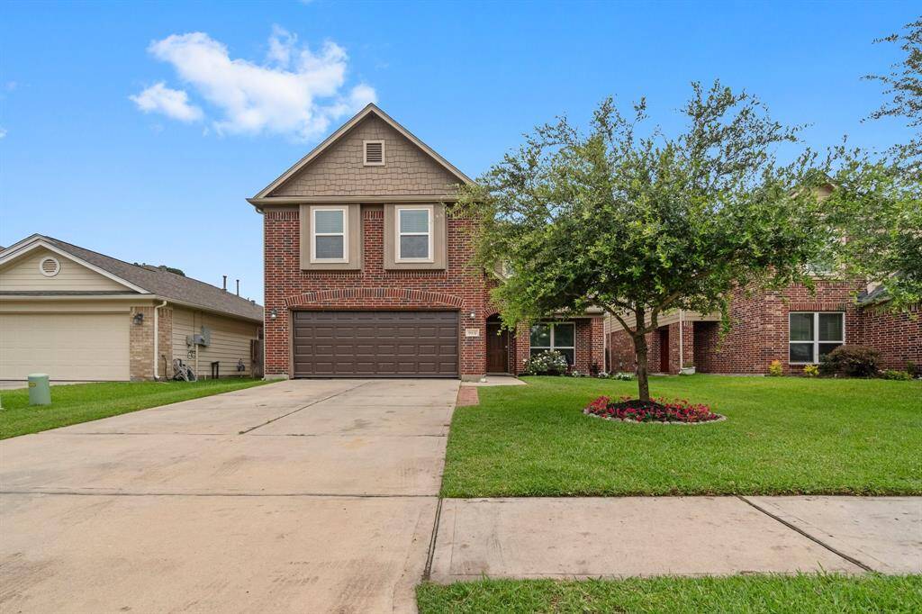 This is a two-story brick home with a gabled roof and a two-car garage. The front yard features a well-maintained lawn, a tree, and a small flower bed. The driveway is spacious, and the house has a welcoming entryway.