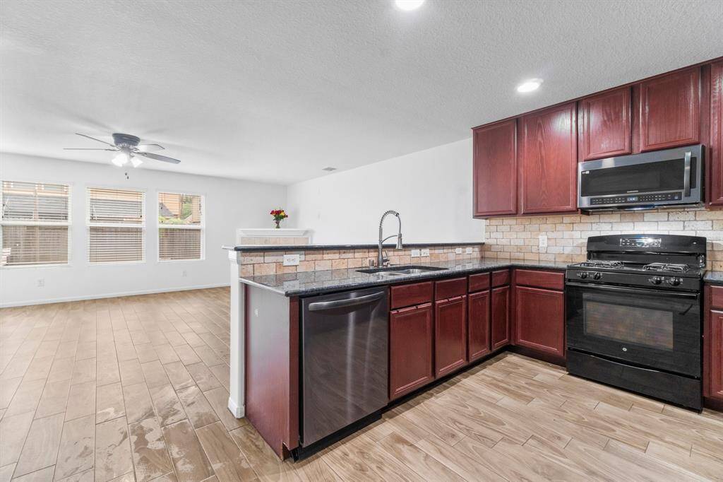 This photo showcases a modern, open-concept kitchen and living area. The kitchen features dark wood cabinets, stainless steel appliances, and a stone backsplash. The living space is bright with large windows and wood-look flooring.