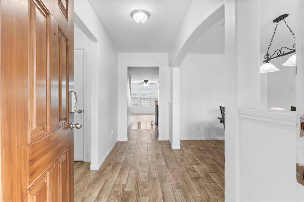 Welcome to a bright and inviting entryway featuring warm wood like tile flooring and a classic door. The hallway leads to a spacious, well-lit living area with large windows.