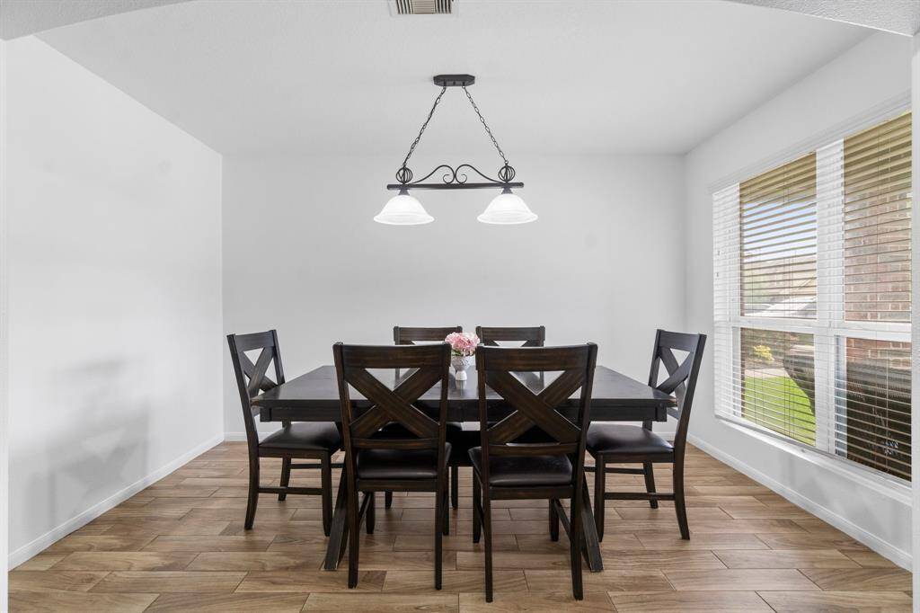 This photo showcases a bright dining area with a modern chandelier.  The room has large windows with blinds, allowing natural light to illuminate the space, and wood-look tile flooring adds warmth.