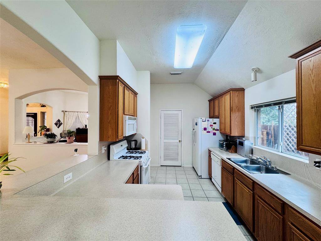 This kitchen features a spacious layout with ample counter space, wooden cabinetry, and modern appliances. It has a large window for natural light and an open view into the adjacent living area.