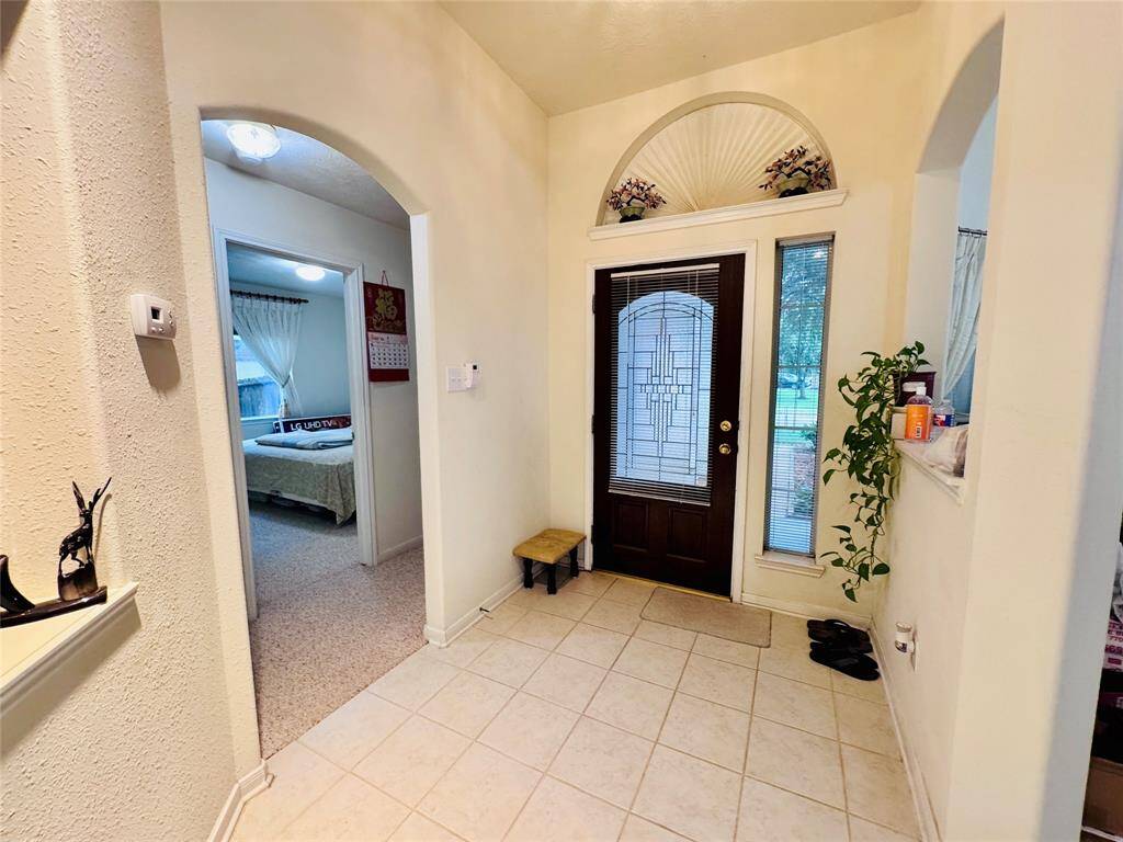 This entryway features a decorative front door with glass panels, tiled flooring, and an arched opening leading to a cozy carpeted room. Natural light enhances the welcoming atmosphere