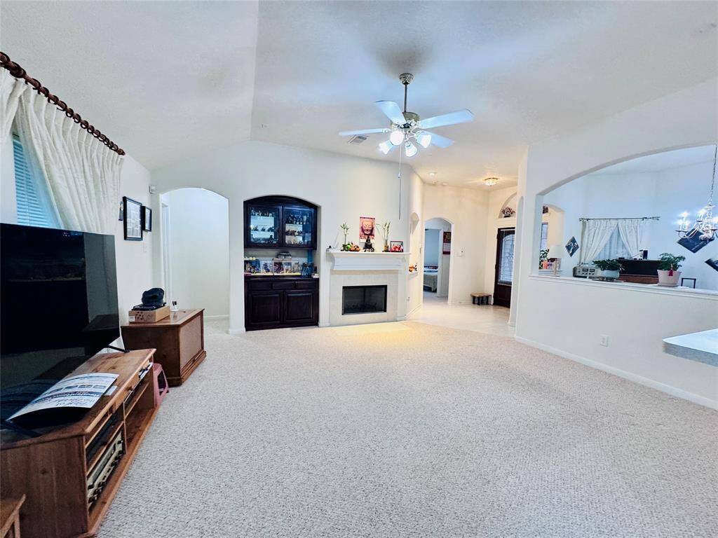 This spacious living room features a light carpet, a cozy fireplace, built-in shelving, and a ceiling fan. There's a large TV area and an open layout leading to the dining space.