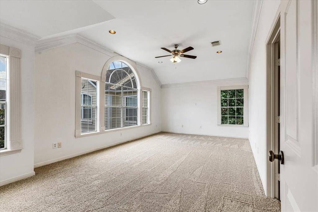 Spacious primary suite with vaulted ceilings, crown molding, and oversized windows—including a striking arched window—that fill the room with natural light and provide a picturesque view of the beautiful mature tree just outside.