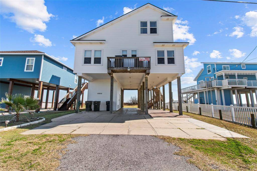 Elevated beach house with a white exterior, featuring a spacious driveway and covered parking underneath. The home has large windows, a small front balcony, and is set in a coastal neighborhood with similar homes. Perfect for enjoying seaside living.