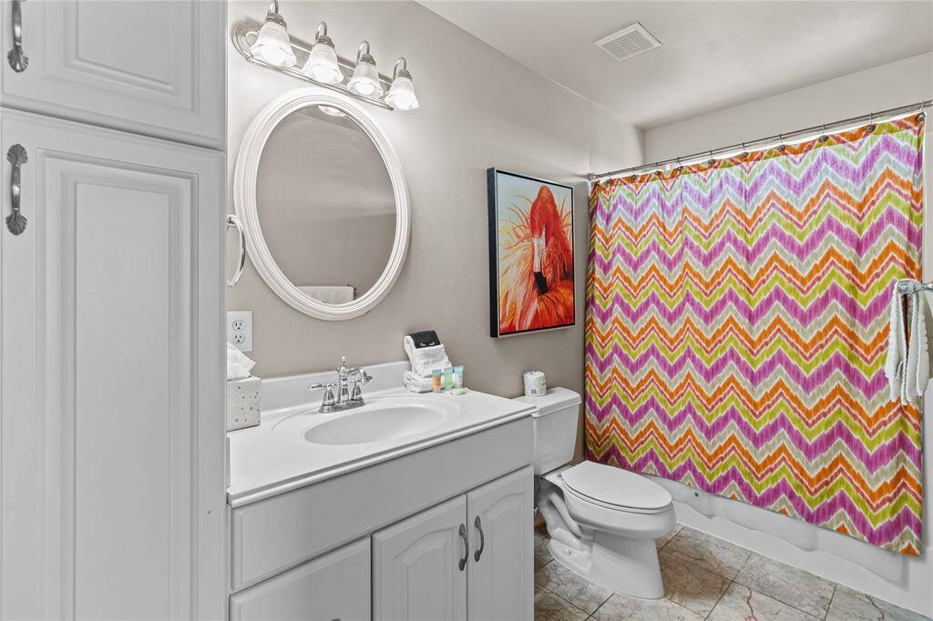Experience a bright, stylish bathroom with a sleek white vanity featuring ample storage, a chic round mirror, and modern lighting. The shower area is enhanced by a vibrant chevron-patterned curtain and tasteful wall art.