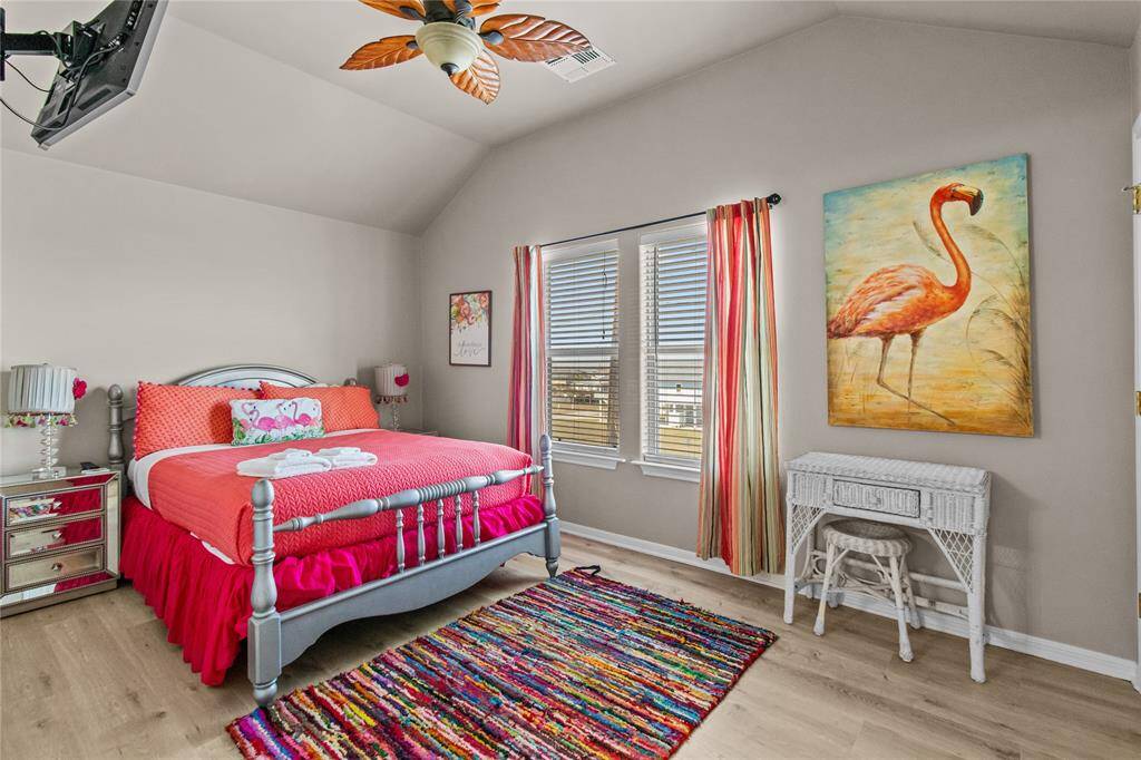 Bright and cheerful bedroom with vaulted ceiling, featuring a colorful bedspread, vibrant area rug, and large flamingo artwork. Natural light pours in through the windows with striped curtains, creating a cozy and inviting atmosphere.