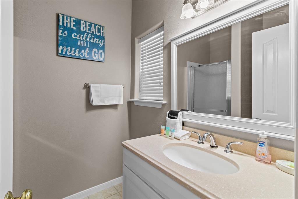 This is a bright, well-lit bathroom featuring a large mirror, a single sink with ample counter space, and a shower. It has a modern feel with neutral tones and a decorative wall sign.