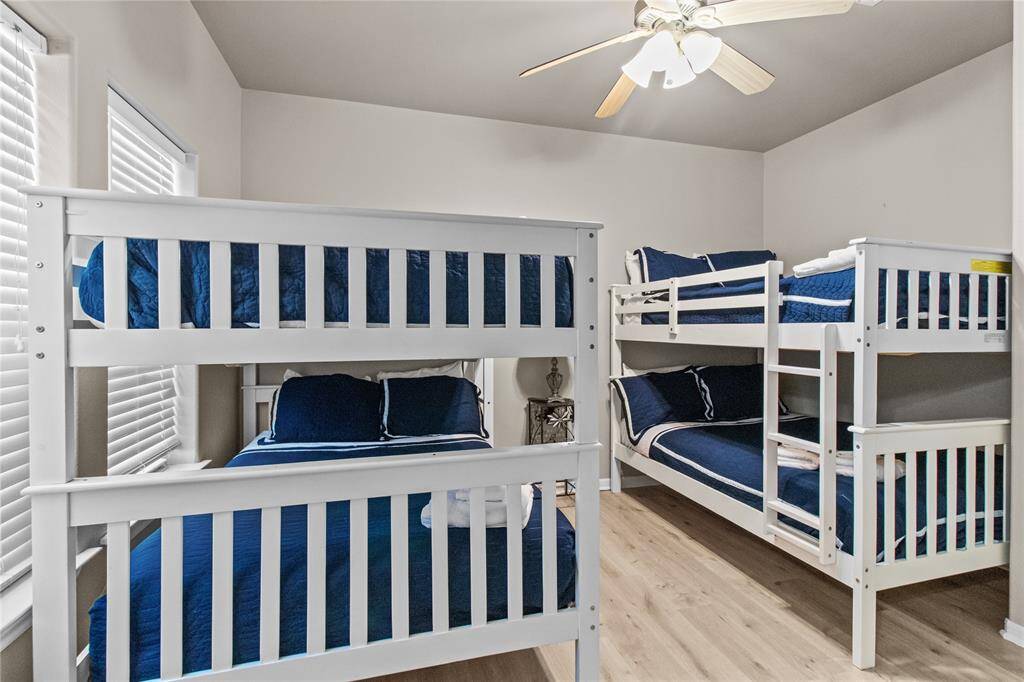 This bedroom features two sets of white bunk beds with blue bedding, ideal for accommodating multiple guests or children. The room is well-lit with natural light from two windows and has a ceiling fan for comfort. The neutral walls and wood flooring add a cozy touch.