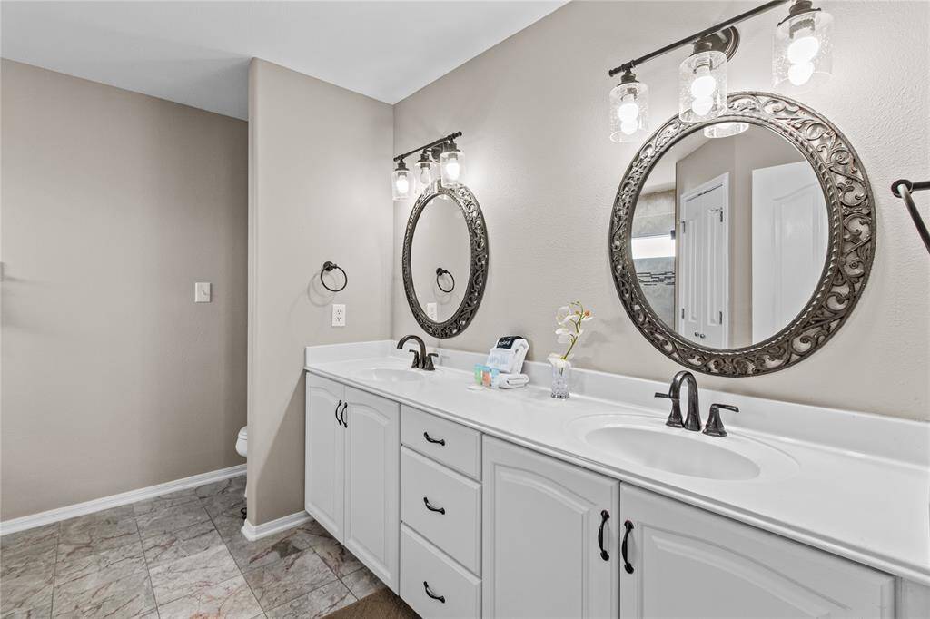 This bathroom features a modern double vanity with elegant white cabinetry and sleek black fixtures. Two ornate mirrors with decorative frames are complemented by stylish overhead lighting. The space is finished with neutral wall colors and a tiled floor, creating a clean and inviting atmosphere.