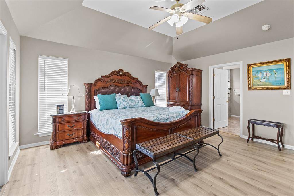 This spacious primary bedroom features elegant wood furniture, including a large bed and matching armoire. It has a vaulted ceiling with a fan, light wood flooring, and two tall windows providing natural light. An en-suite bathroom is accessible through a door, enhancing convenience.