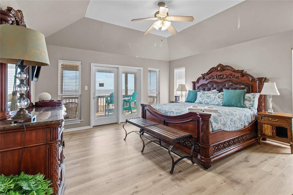 This spacious bedroom features elegant wooden furniture, including a large, intricately carved bed. Natural light streams through multiple windows and French doors, which open to a balcony with a view. The room has a vaulted inset ceiling with a ceiling fan and light wood flooring, creating a bright and airy atmosphere.