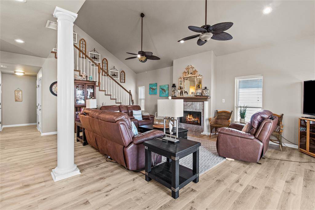 Spacious living room with high ceilings, featuring a cozy fireplace and elegant wooden flooring. The room is furnished with comfortable leather seating and has a stylish staircase leading to the upper level. Ceiling fans and ample natural light enhance the inviting atmosphere.