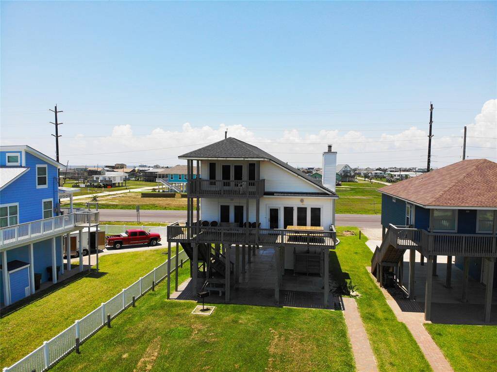 This photo shows a two-story home elevated on stilts, featuring a spacious balcony and a covered area below. Offers a golf cart and/or beach toy storage area.  It is situated between two other houses, with a fenced yard and a clear view of the surrounding area.