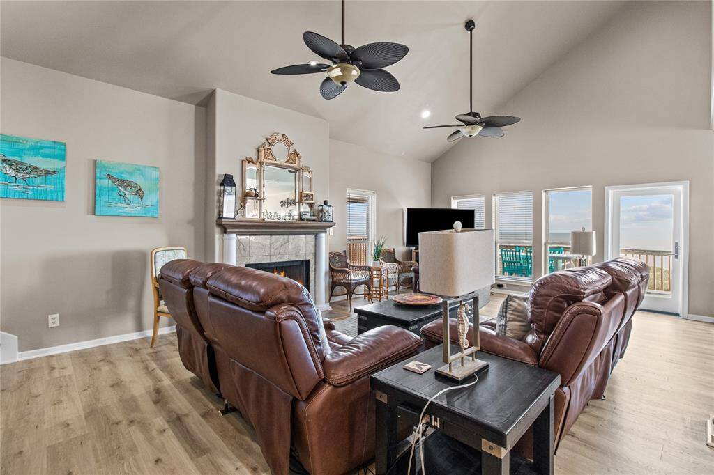 This spacious living room features a high ceiling with modern fans, a cozy fireplace, and elegant wood flooring. It includes comfortable leather seating, a stylish staircase, and ample natural light, creating a warm and inviting atmosphere.