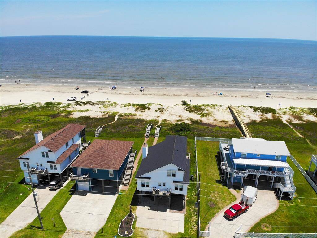 This photo shows a beachfront neighborhood with elevated homes featuring ocean views. The property is near a wide sandy beach, offering easy access to the ocean. The area includes a main road for convenient travel, and the homes have spacious yards. Ideal for those seeking a coastal lifestyle.