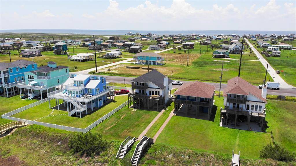 This photo shows a coastal neighborhood with elevated homes, ideal for beach living. The houses are situated on spacious lots with green lawns, and a road runs parallel to the shoreline, offering easy access to the beach. The area is open and scenic, with a view of the ocean in the background.