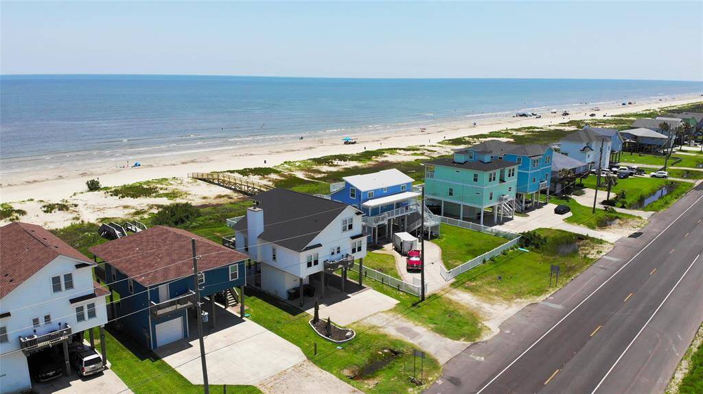 This coastal photo showcases a row of colorful beachfront homes with direct access to a sandy beach and expansive ocean views. The homes are elevated, featuring driveways and lush green lawns, ideal for a serene seaside lifestyle.