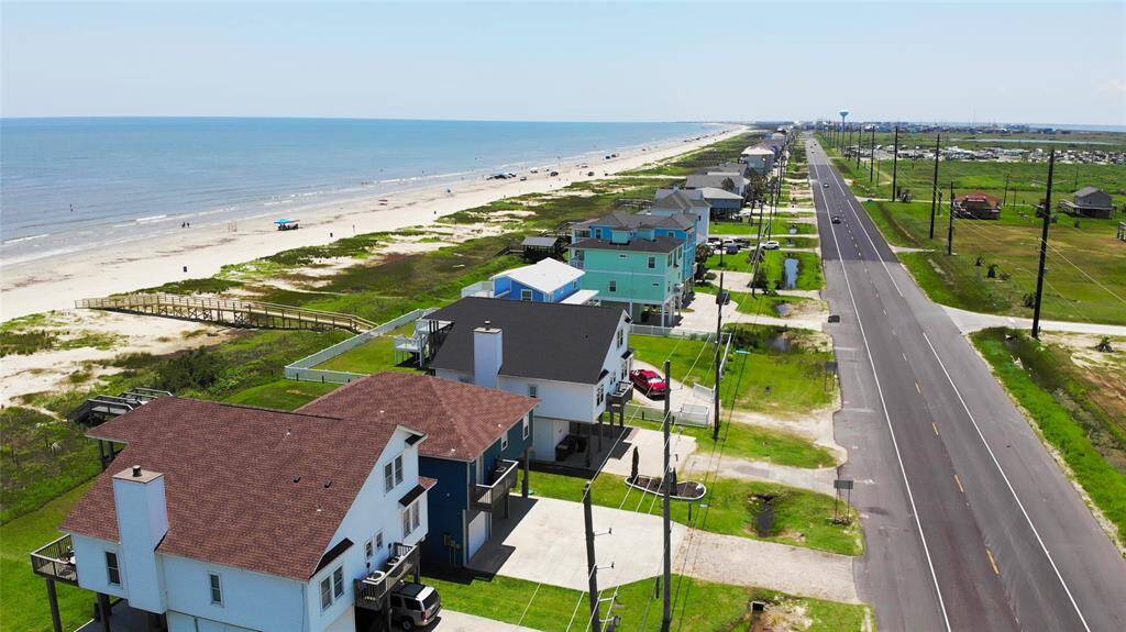 This aerial photo shows a row of elevated beach houses with spacious decks and fenced yards. The homes are located near a road and surrounded by a mix of grassy areas and neighboring properties, offering a coastal community setting.