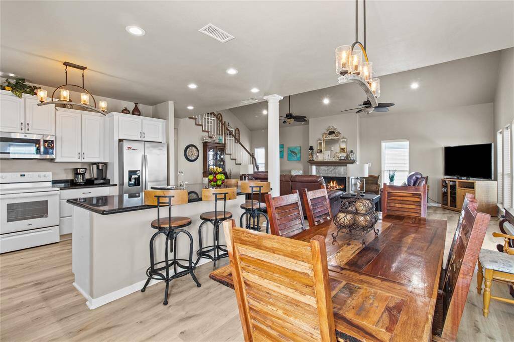 This open-concept space features a modern kitchen with white cabinets, stainless steel appliances, and a breakfast bar with seating. It flows seamlessly into a cozy living area with a fireplace and a dining space with a rustic wooden table. Natural light enhances the airy feel, complemented by neutral tones and elegant lighting fixtures.