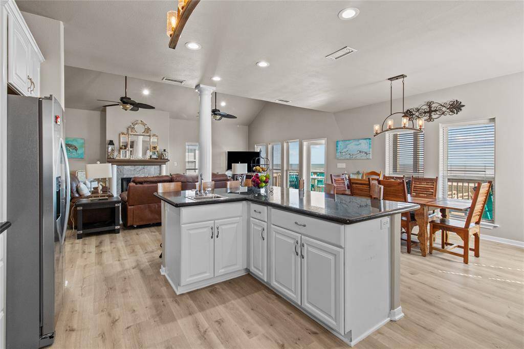 This kitchen features modern white cabinetry with glass accents, granite countertops, and stainless steel appliances. It includes a spacious island with a double sink and ample storage. The area is well-lit with stylish pendant lights and has light wood flooring that adds warmth to the space.