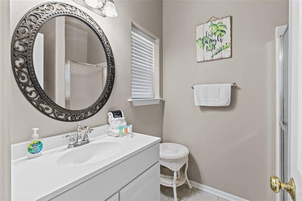 This bathroom features a clean, modern design with a white vanity and sink, a decorative round mirror, and a neutral color scheme. It has a window for natural light, a towel rack, and a decorative wall piece, creating a fresh and welcoming space.